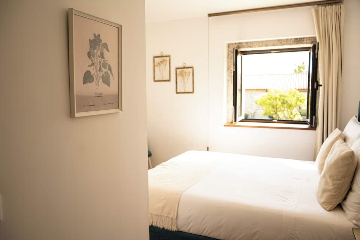 A bedroom with a bed, a window, and framed botanical prints on the wall.