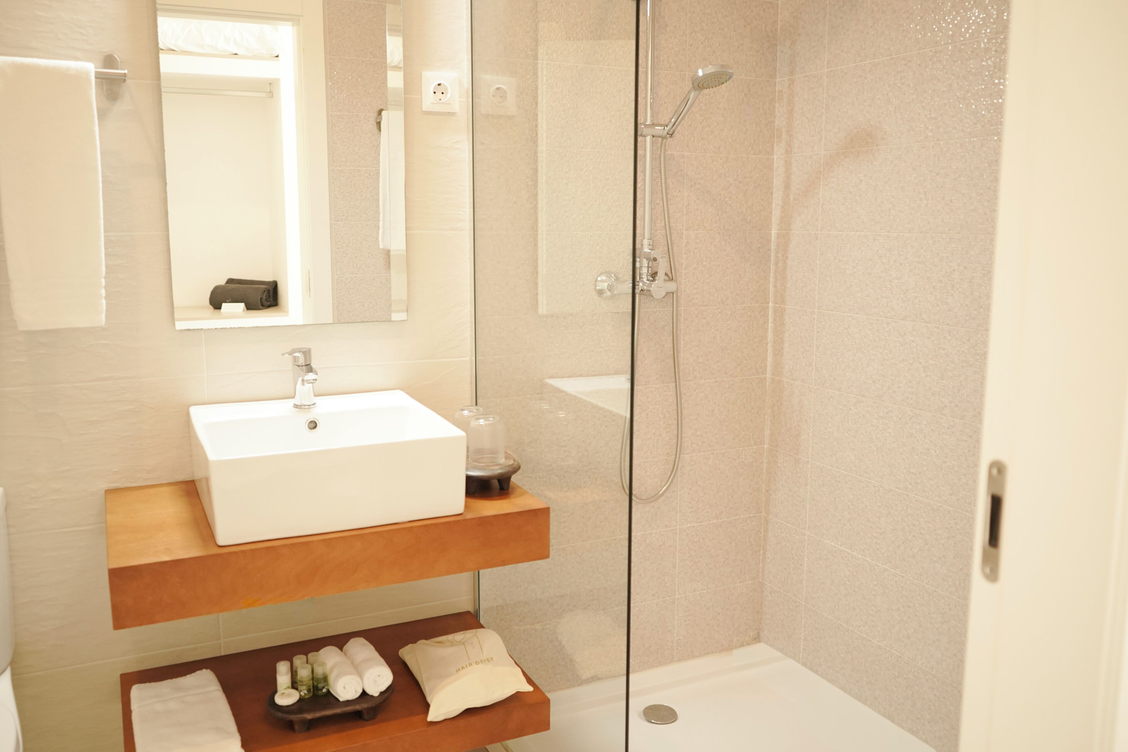 Un baño moderno con cabina de ducha de vidrio, un lavabo blanco y un estante de madera con artículos de aseo.