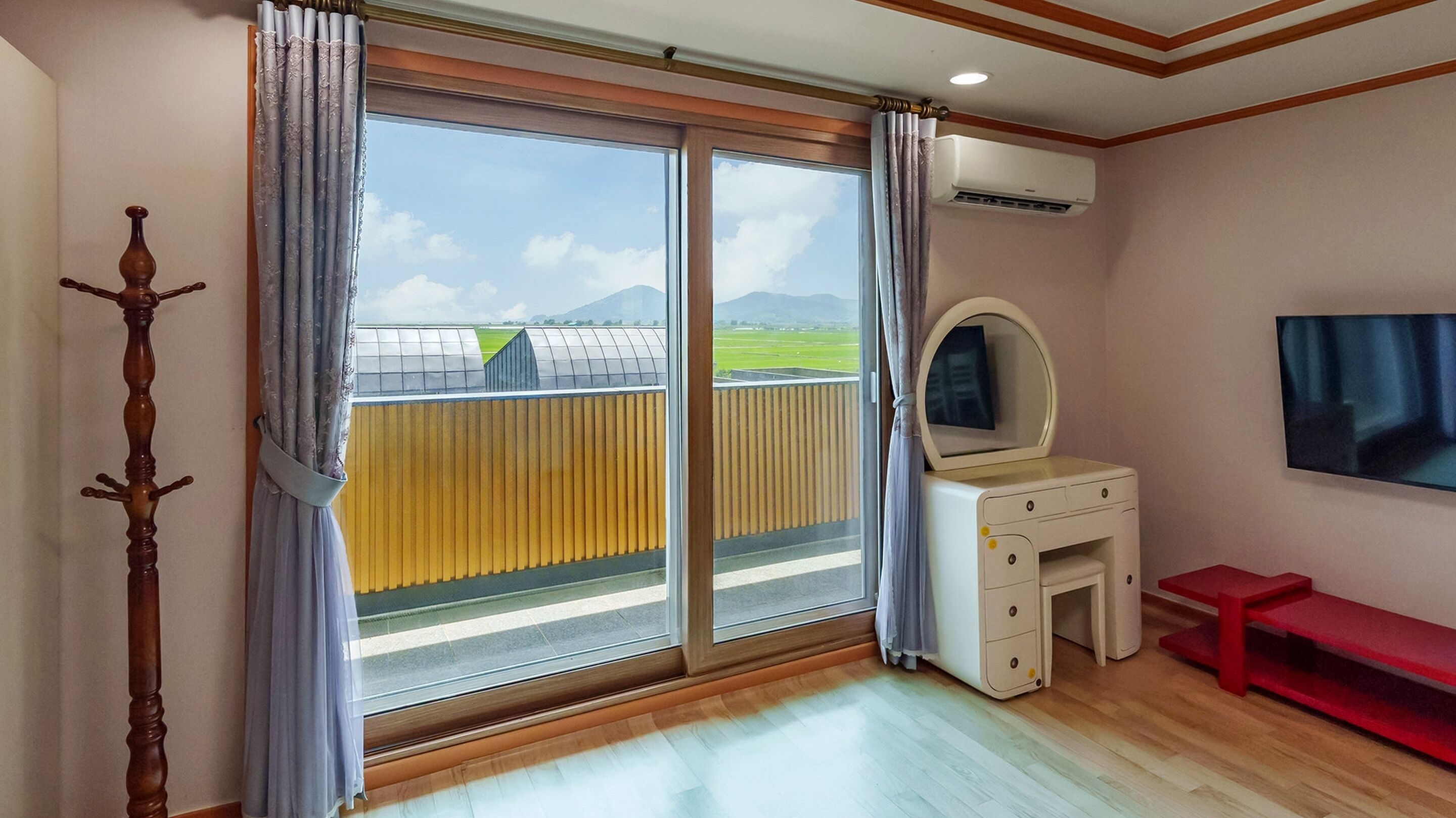 A bedroom with a sliding glass door to a balcony, a vanity with a mirror, a red bench, and an air conditioning unit on the wall.