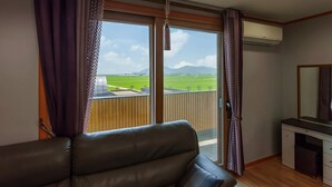 Una habitación con sofá de cuero, un escritorio pequeño y un amplio ventanal corredero que da a un balcón con vistas a un campo verde y montañas.