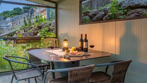Una mesa de comedor con una vela, botellas de vino y una copa de vino en un balcón con sillas de mimbre y vistas a un jardín rocoso.
