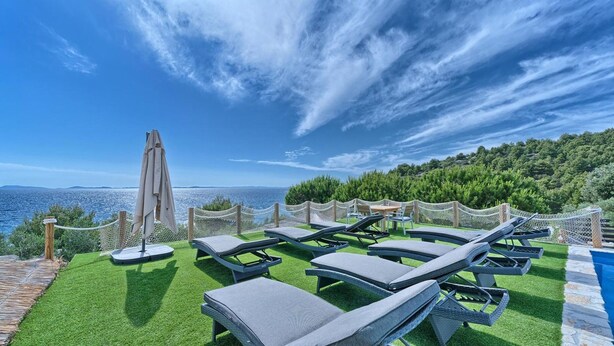 Zona junto a la piscina con sillones de descanso y sombrillas, con vistas a un cuerpo de agua y una ladera boscosa.
