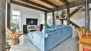 Un salon moderne avec un grand canapé d’angle bleu, des poutres en bois au plafond, une table à manger avec des chaises et un escalier.