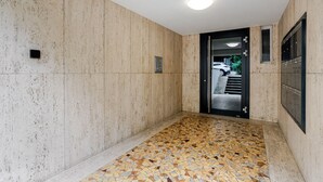 Um corredor estreito com piso ladrilhado, uma porta que dá para área externa e uma luminária fixada na parede.