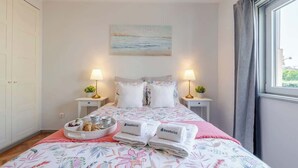 Un llit ben arropat amb roba de llit floral, dues làmpades de nit, un quadre de paisatge mariner i una finestra amb cortines.