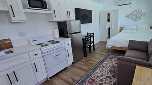 A compact kitchen with white cabinets, a microwave, a refrigerator, and a stove. There is a bed with white bedding and a brown couch. A patterned rug is on the floor.