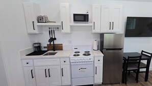 A compact kitchen with white cabinets, a microwave, a stainless steel refrigerator, a stove, and a dining area with a chair.