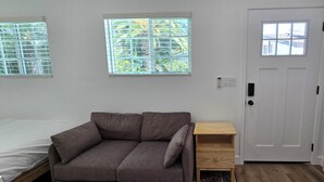 A room with a bed, a brown sofa, a small wooden nightstand, a white door, and two windows with blinds.