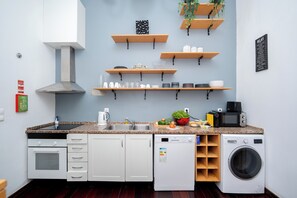 A compact kitchen with a microwave, sink, oven, and dishwasher. There are wooden shelves above the counter holding various kitchen items. - Palma Rooms Central Lisbon (Lisbon)