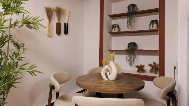 Un comedor con una mesa redonda de madera, cuatro sillas, una planta en maceta y estantes colgantes con plantas y adornos.