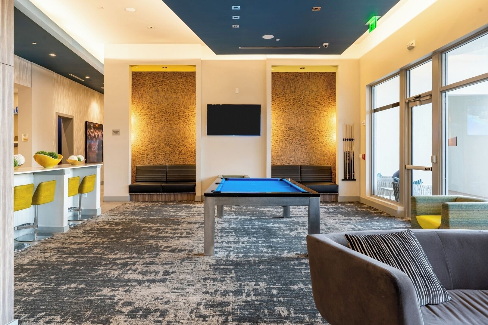 Een moderne biljarttafel in een ruimte, met een blauwe viltkleed, omringd door een vloerkleed en een zithoek met een bank en stoelen.