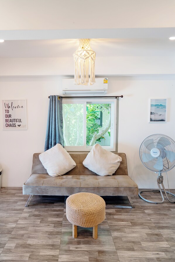 Una habitación con sofá, dos almohadas, un reposapiés tejido, una ventana con cortinas, un ventilador de pared y un letrero.