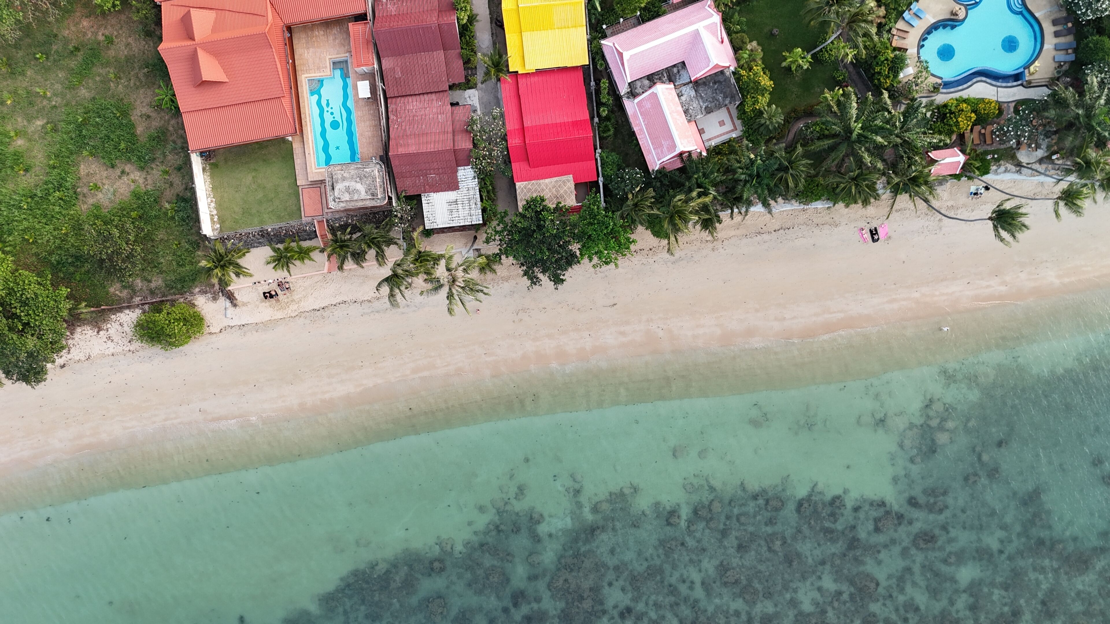 Vista aérea de una propiedad frente al mar con edificios coloridos, piscina y playa de arena.