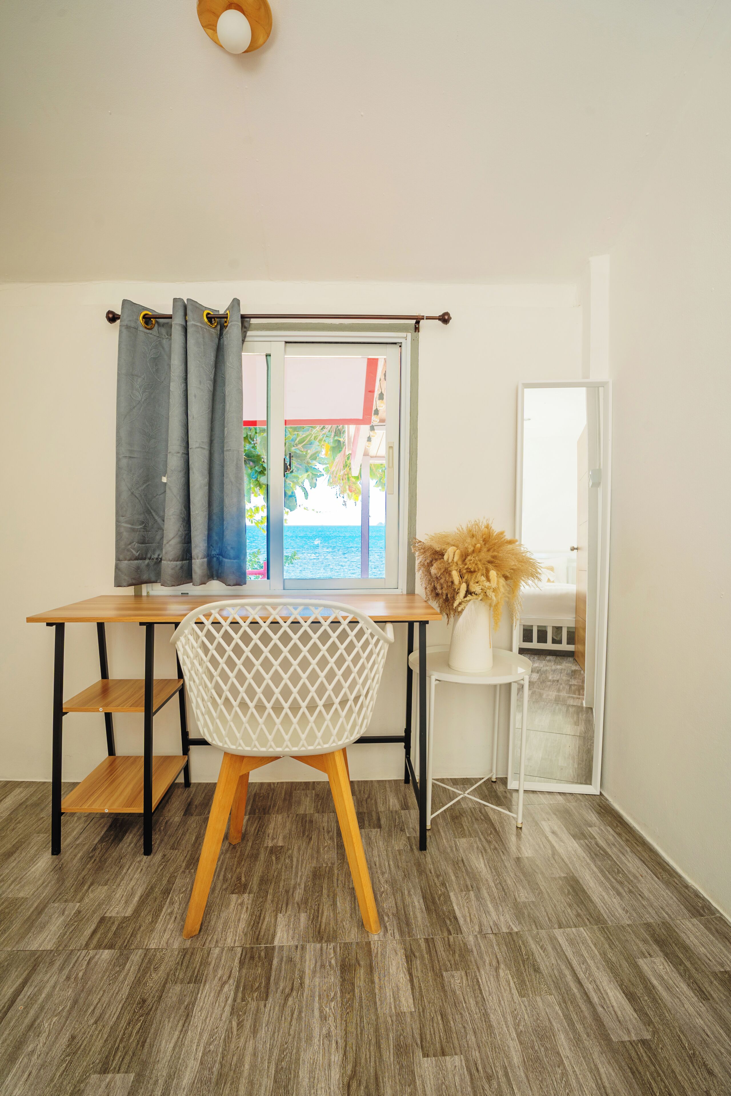 A room with a wooden desk, a white chair, a small white table with a vase, and a window with curtains.