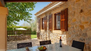 An outdoor patio with a table set for dining, stone walls, wooden shutters, and a view of a garden. - ES CLAPER - Beautiful estate with private pool and free Wi-Fi in Artà (Artà)