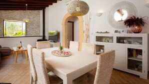 A dining area with a white table, wicker chairs, a hanging light fixture, and a white cabinet with shelves. - ES CLAPER - Beautiful estate with private pool and free Wi-Fi in Artà (Artà)