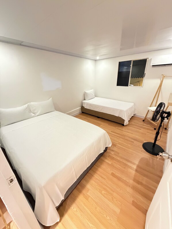 A room with a large bed, a smaller bed, a standing fan, and a window with a view.
