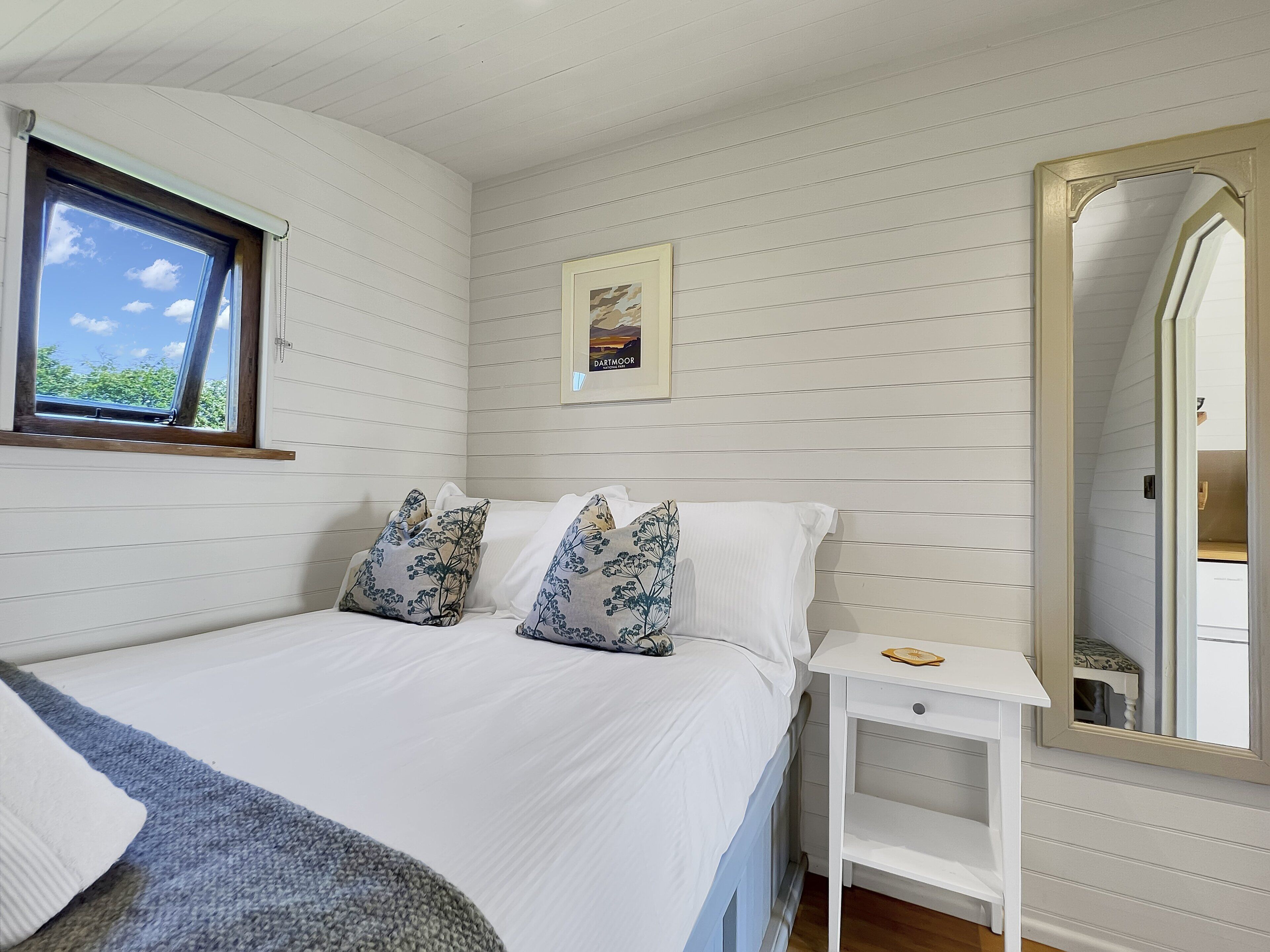 A bedroom with a bed, pillows, a bedside table, a framed picture, a window, and a mirror.
