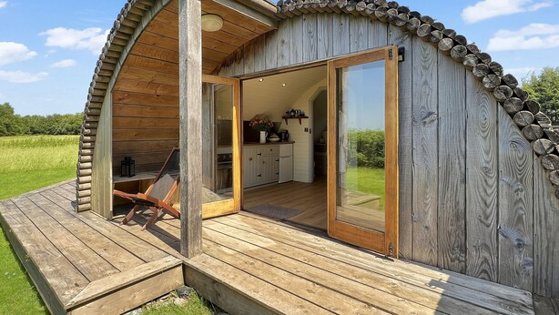 Een houten vakantiehuis met een terras, een glazen deur naar een keuken en uitzicht op een groen veld.