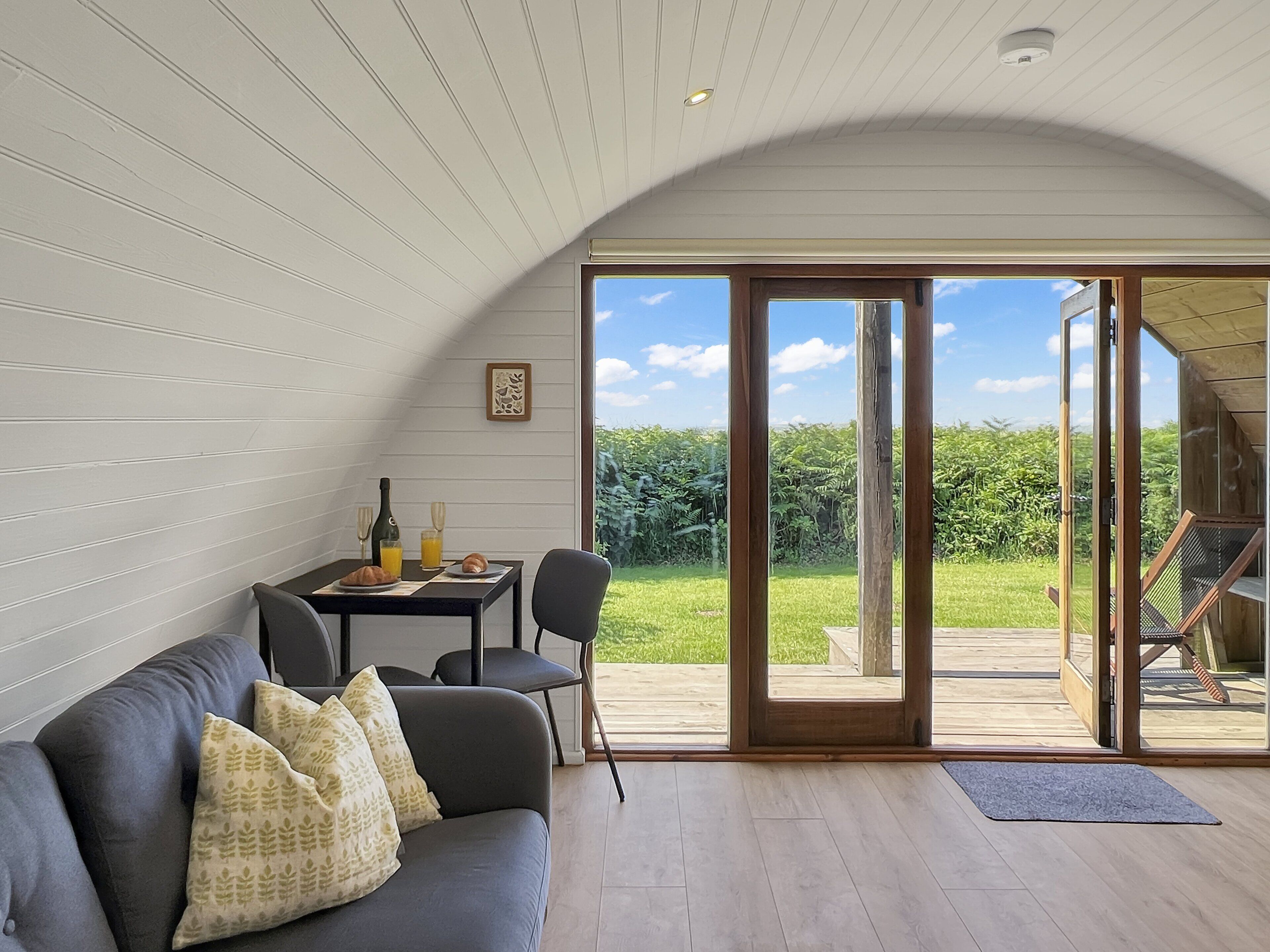 A modern interior with a curved ceiling, a dining area with a table and chairs, a sofa with patterned cushions, and a large sliding glass door leading to an outdoor deck.