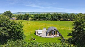 Een rustiek huis met rieten dak, een terras met tuinmeubels en een uitzicht op een groen veld en heuvels op de achtergrond.