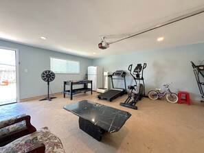 Une salle de sport à domicile équipée d’appareils d’exercice, d’un ventilateur, d’un réfrigérateur et d’une fenêtre avec des stores.
