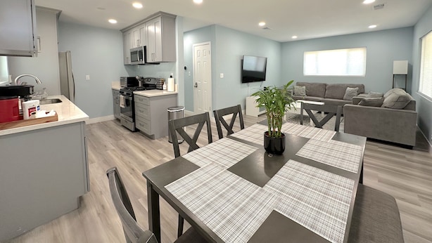 A modern kitchen with grey cabinets, a microwave, and a stove. There is a dining area with a table and chairs, and a living room with a sofa and a TV. - Stay Near Disneyland – Clean, Spacious Anaheim Home for Families & Friends (Anaheim)
