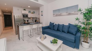 Ein modernes Wohnzimmer mit einem blauen Sofa, einem weißen Couchtisch und einer Küchenzeile mit weißen Schränken und einem Kühlschrank.