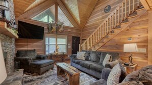 A log cabin interior with a stone fireplace, a wooden staircase, and a cozy living area with a sofa and coffee table.