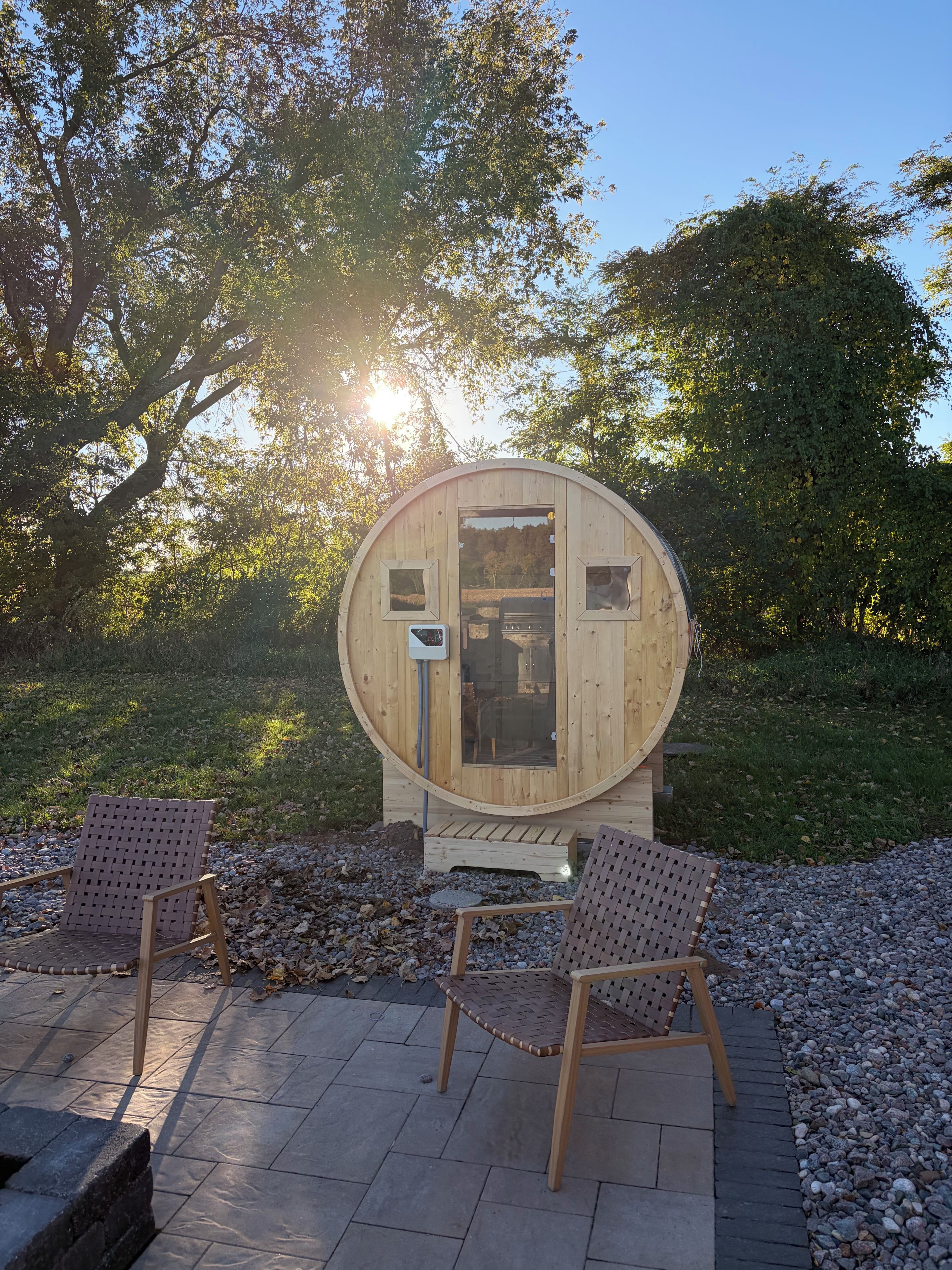 Estructura de sauna de madera con dos sillas en el frente, rodeada de árboles y grava.