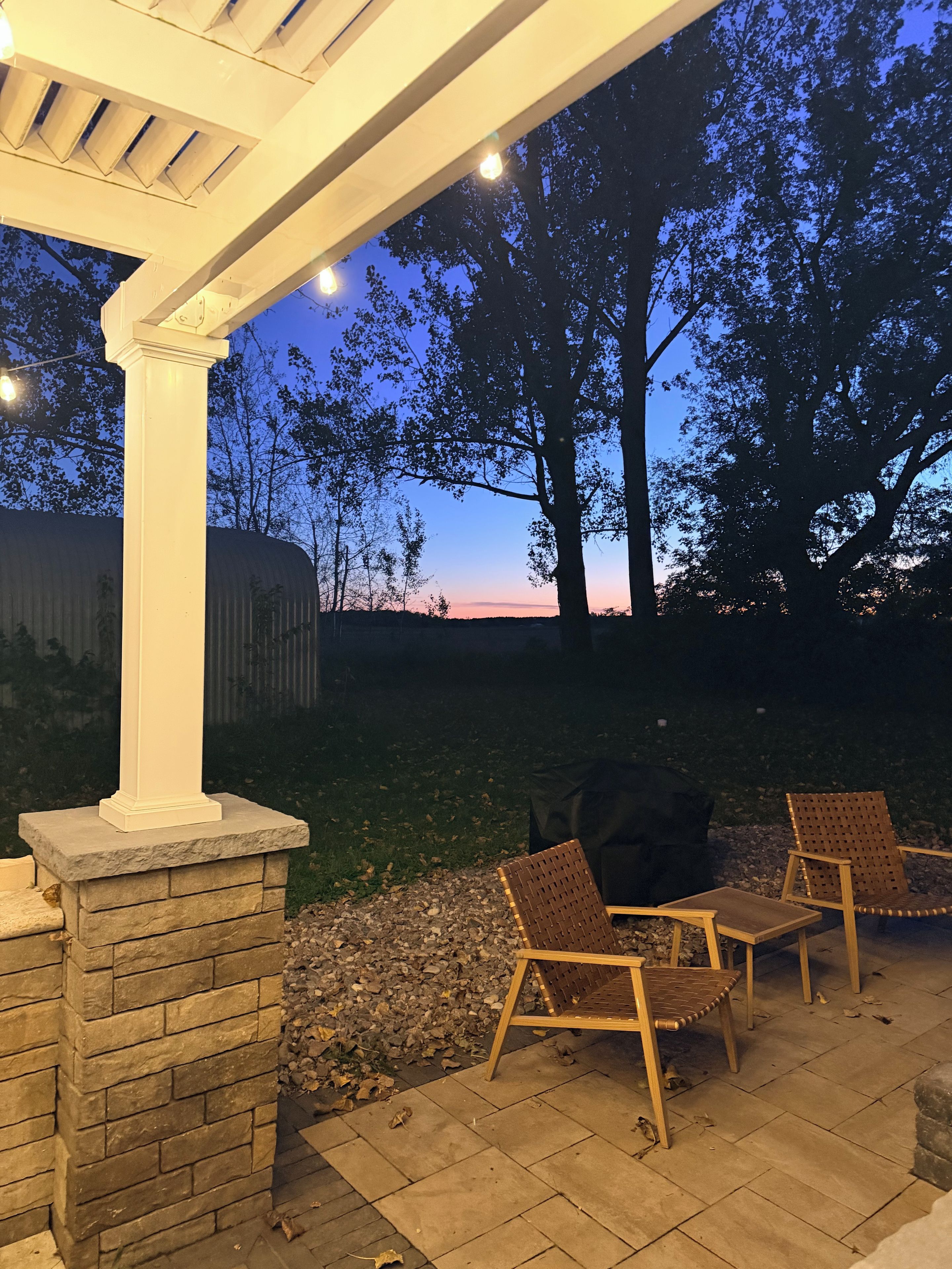An outdoor patio with a pergola, stone pillar, and two wicker chairs.