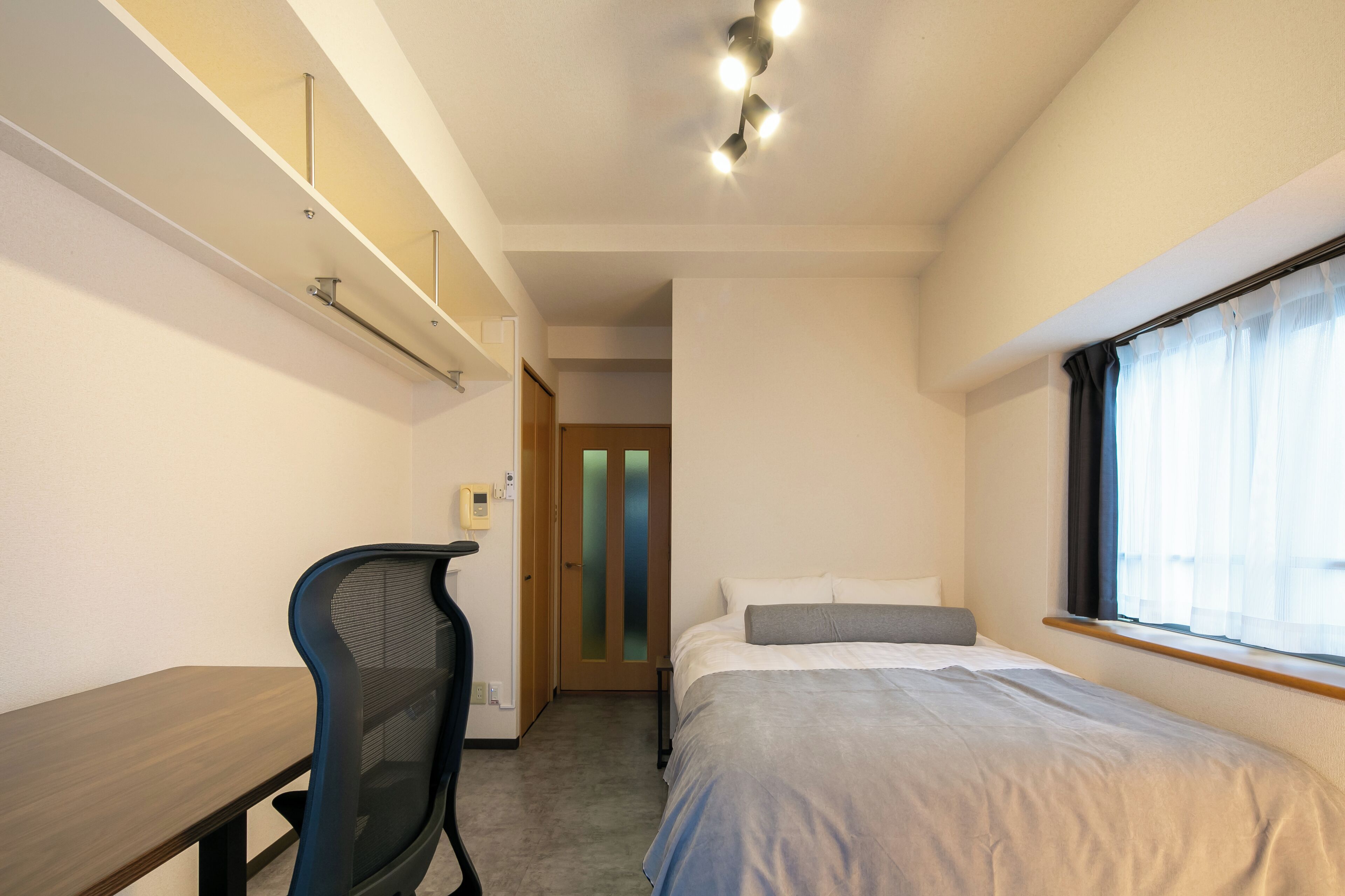 A compact bedroom with a single bed, a desk, and a chair. There is a window with sheer curtains and a door with frosted glass panels.