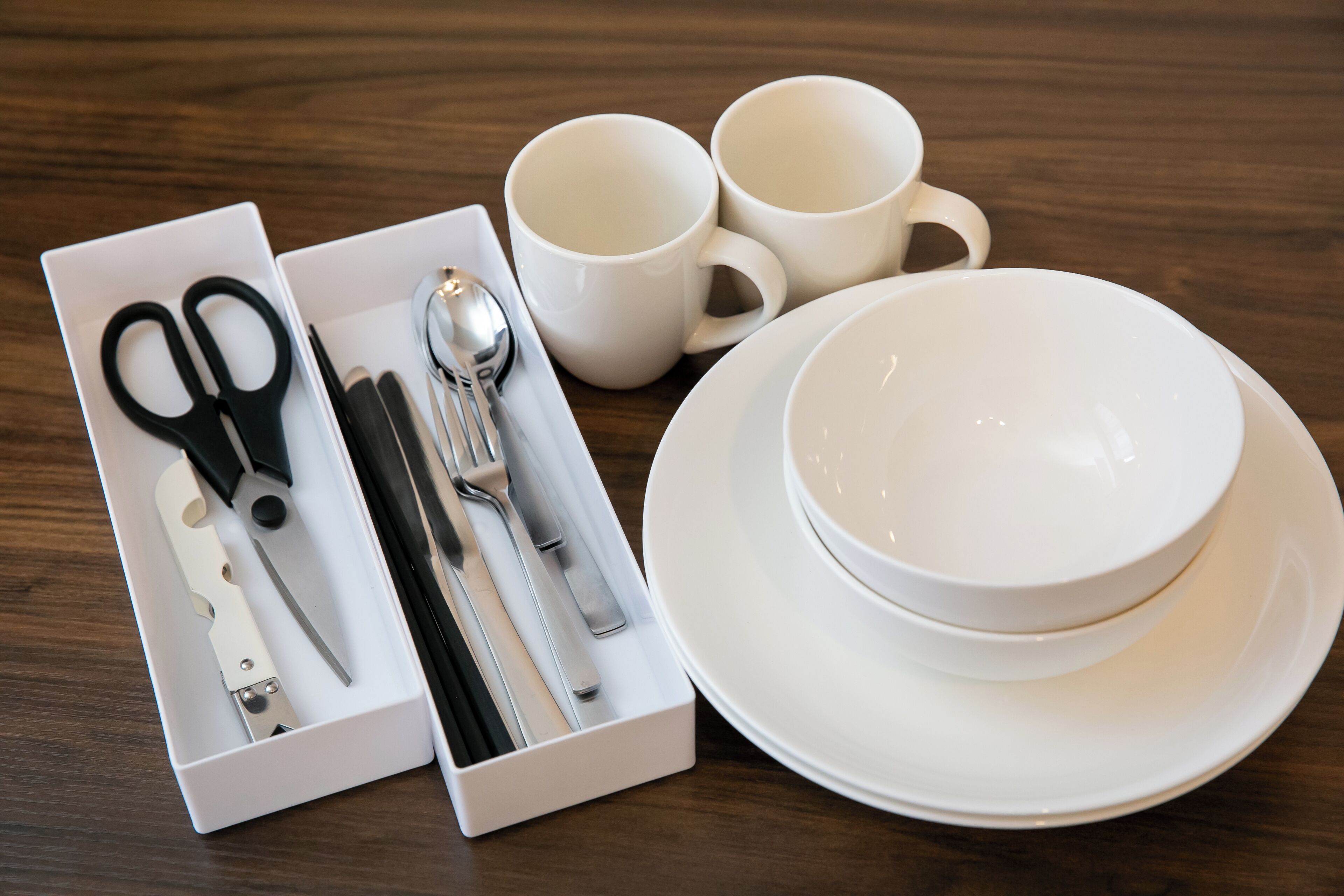 Un juego de cubiertos blancos, que incluye un cuchillo, una cuchara y un tenedor, junto con dos tazas blancas y un bol blanco sobre una mesa de madera.