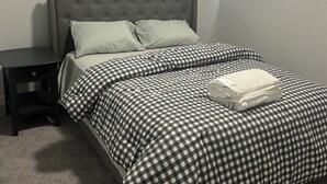 Un lit bien fait, avec une couvre-lit à carreaux, une tête de lit rembourrée de couleur grise et une petite table de chevet dotée d’un tiroir.