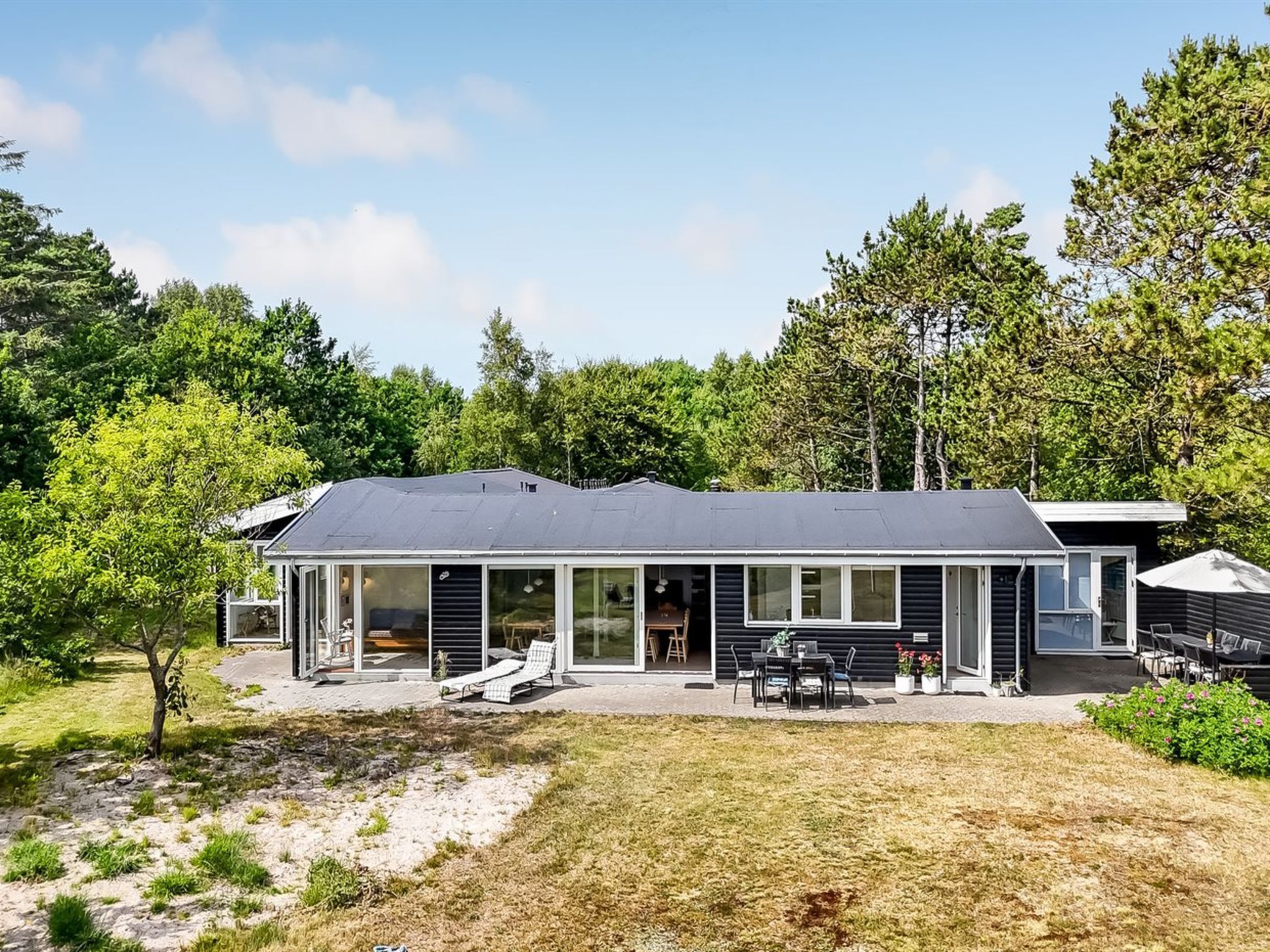 A modern house with a large glass front, a patio with outdoor furniture, and a surrounding wooded area.