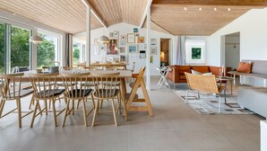 Un interior espacioso y moderno con vigas de madera en el techo, un comedor con mesas y sillas de madera, una sala de estar con sofá y sillones, y obras de arte en las paredes.