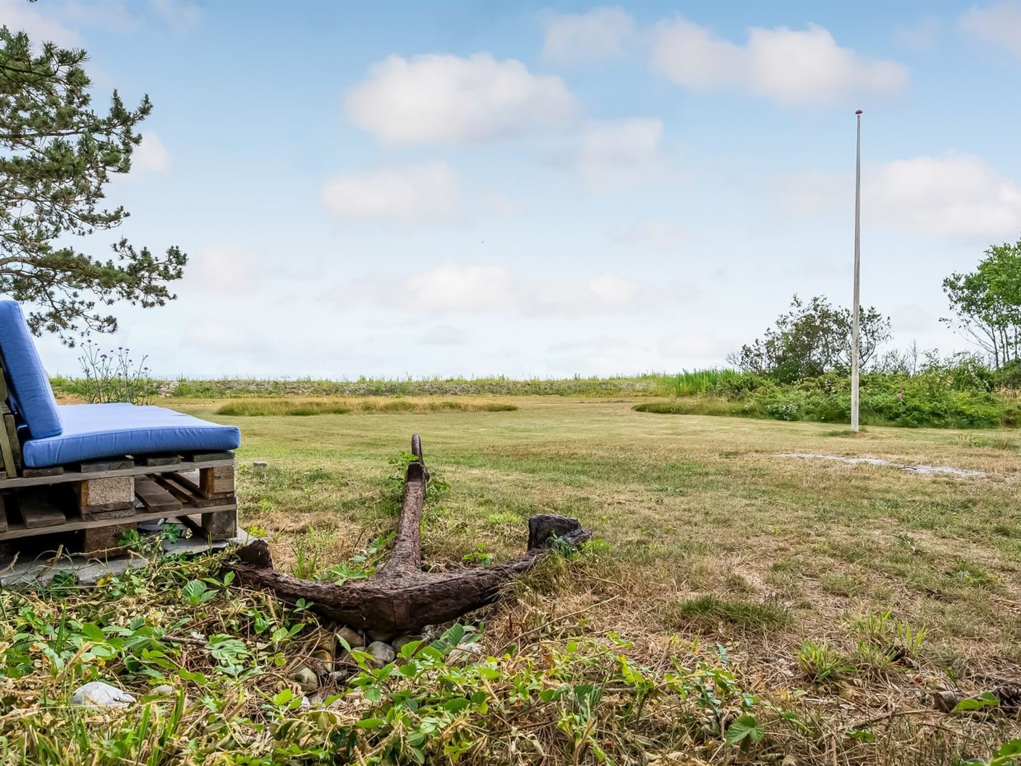 Une terrasse en bois avec une chaise rembourrée bleue, une sculpture d’ancre rustique et un ciel dégagé.