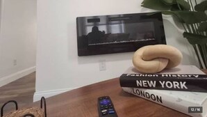 Une pièce avec un téléviseur à écran plat fixé au mur, un tableau encadré, une plante en pot, une pile de livres et une télécommande posée sur une table en bois.