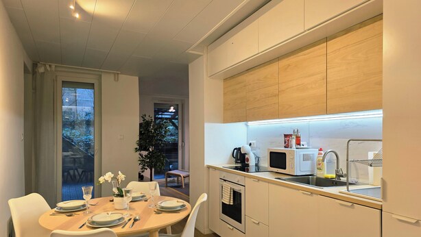 A modern kitchen with white cabinets, a microwave, a kettle, and a sink. There is a dining area with a table set for two.