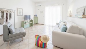 Un salon avec un canapé gris, un canapé blanc, un sac à rayures colorées, un chapeau de paille et une paire de tongs posées sur le sol.