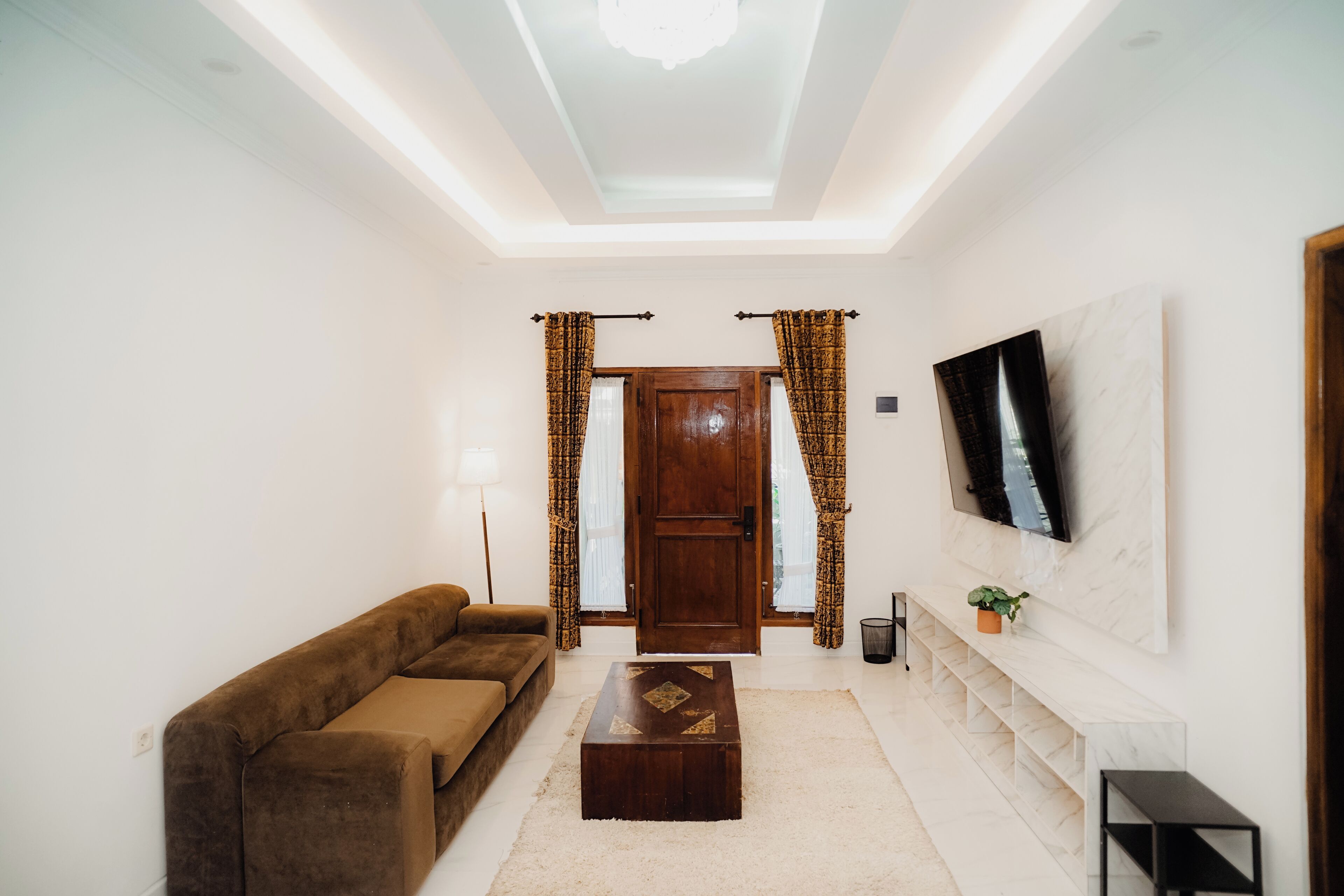 A living room with a brown sofa, a wooden coffee table, a TV mounted on the wall, and a door with curtains.