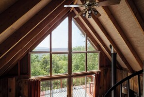 Une pièce dotée d’une grande fenêtre, de poutres en bois, d’un ventilateur de plafond et offrant une vue sur un paysage boisé.