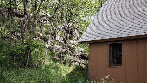 A small wooden cabin with a sloped roof, surrounded by dense forest and rocky terrain.