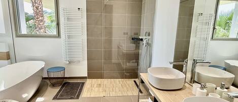 A modern bathroom with a freestanding bathtub, two sinks, a shower, and a large window with a view of greenery.