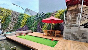 Une table de billard entourée d’une terrasse en bois, un parasol rouge et un mur de pierre agrémenté de verdure.