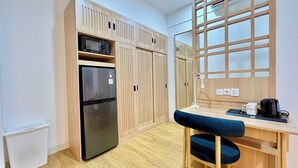 Une cuisine compacte équipée d’un réfrigérateur, d’un four à micro-ondes et de meubles de rangement en bois. On y trouve un petit bureau avec une chaise, une lampe et une machine à café.