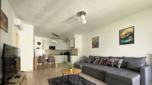 Un salon moderne avec un canapé gris, une table basse en bois et un tapis noir à poils longs. On y trouve également un coin repas avec une table et des chaises en bois, et une cuisine à l’arrière-plan.