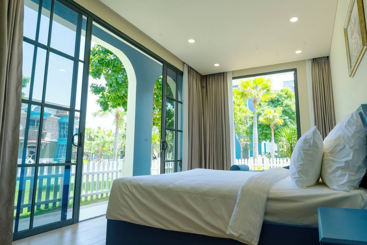 A hotel room with a large bed, a nightstand, and a view of a garden through large windows.