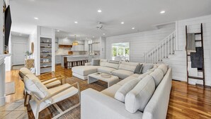 A spacious living area with a large L-shaped sofa, wooden flooring, and a kitchen in the background. - 7BR Estate · One Block to Ocean · Sleeps 22 (Falmouth)