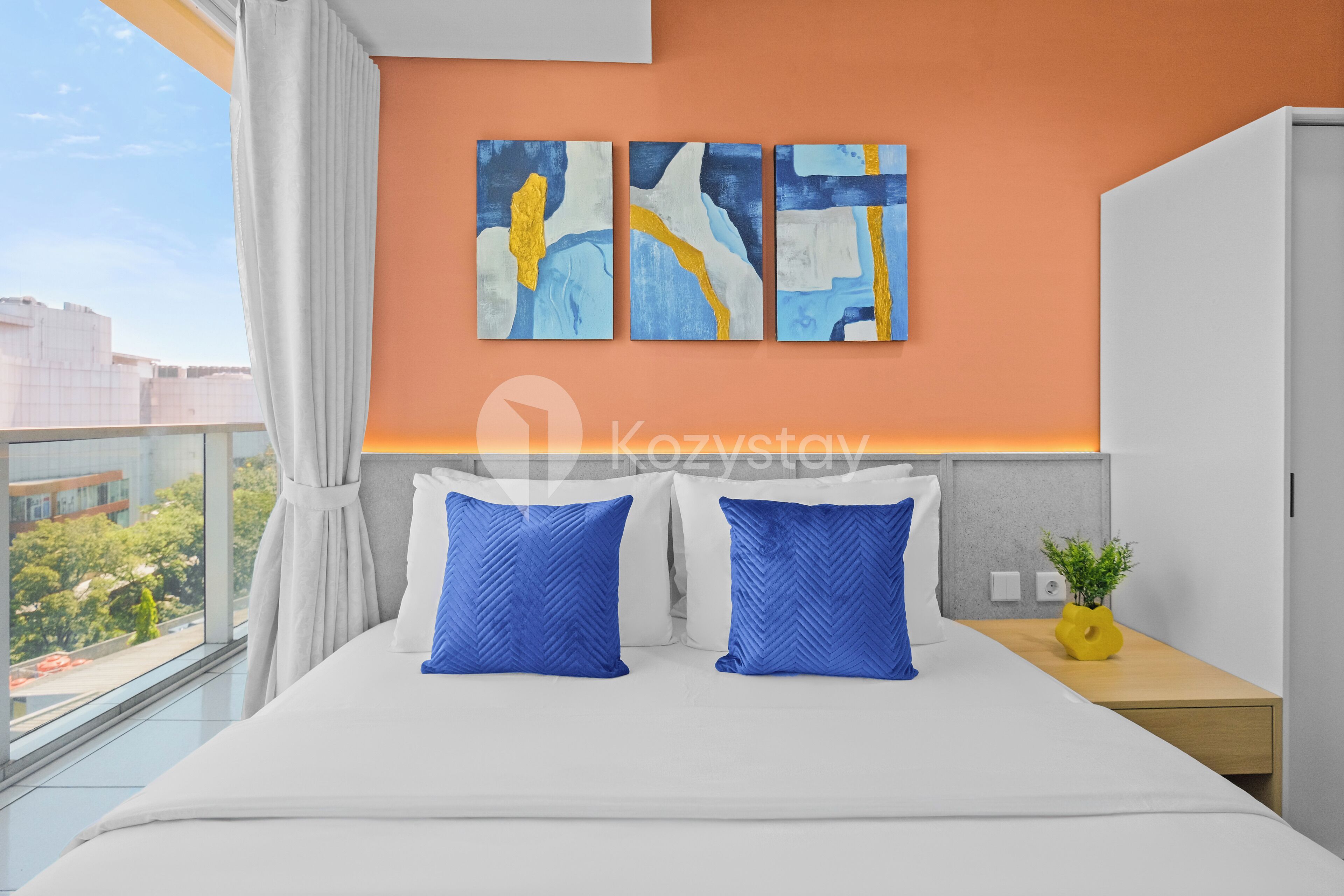 Quarto de hotel moderno com uma cama grande, duas almofadas azuis, uma mesa de cabeceira com uma planta e arte abstrata na parede.